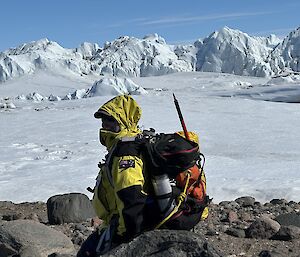 A person in field survival gear and pack in the foreground with massive peaks of snow and ice across the not too distant horizon
