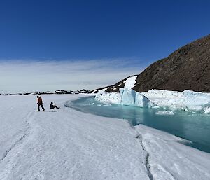 It looks like a snow beach with the snow running down to a turquoise edge of water or maybe ice. Two people are enjoying the view