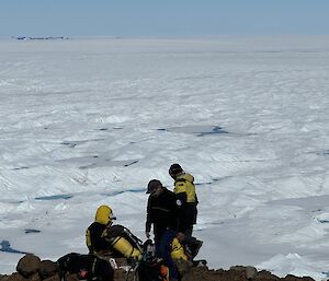 Three people in the foreground dwarfed by a massive and bumpy icefield stretching away to the distant horizon where a rock outcrop appears tiny