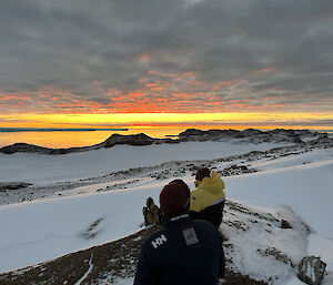 A beautiful sunset in shades of yellow and orange across a foreground of snowy rocks and two people watching. The tongue of a glacier can be made out as a thin line on the horizon.