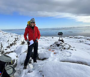 A smiling man in a red jacket stands in the snow with a camera on a tripod behind him