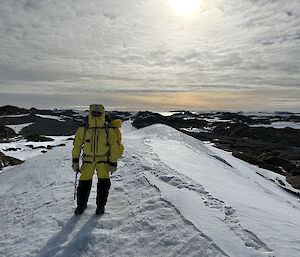 An unidentifiable person in a yellow AAD suit stands on a snow ridge with a rocky peninsula in the background