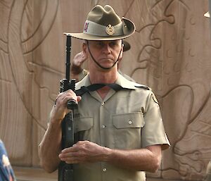 A man in a khaki soldiers uniform including a slouch hat stands to attention with his rifle by his side