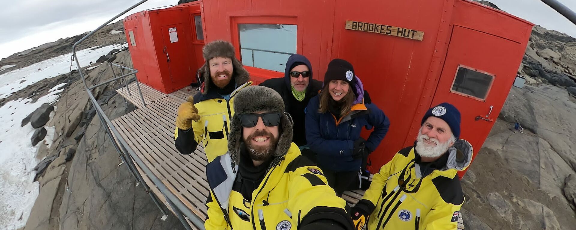 A group of five smiling people standing out the front of a red field hut with a sign on the front, "Brookes Hut." The hut is sitting on a rocky outcrop and snow can be seen surrounding it.