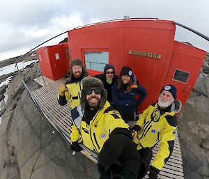 A group of five smiling people standing out the front of a red field hut with a sign on the front, "Brookes Hut." The hut is sitting on a rocky outcrop and snow can be seen surrounding it.