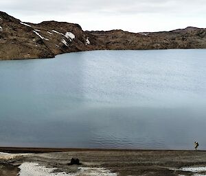 A long-distance shot of a man hiking along the shores of a large lake surrounded by rocky hills