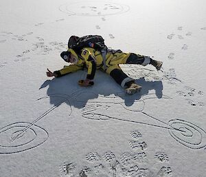 A smiling man lies on his side in the snow and pretends to ride a motorbike that has been drawn into the snow.