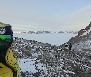 Three people are on an icy, rocky hillside with ice and mountains in the distance