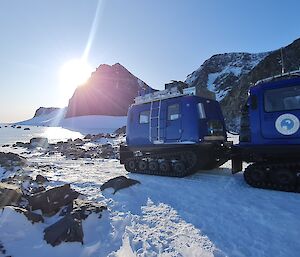 The sun is rising over a mountain peak with a blue articulated snow vehicle in the foreground