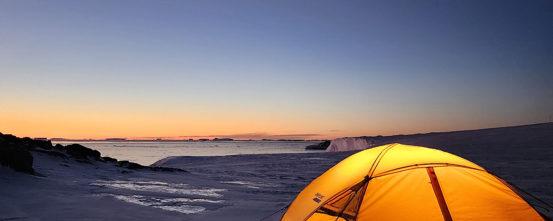 A small yellow tent with a glowing light inside is located in the foreground of the picture. It is set up on snow covered ground and in the distance is a glimpse of the water, and distant icebergs. The sky is glowing orange and blue in the twilight.