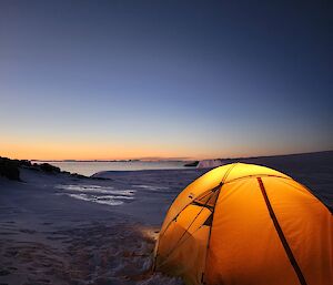 A small yellow tent with a glowing light inside is located in the foreground of the picture. It is set up on snow covered ground and in the distance is a glimpse of the water, and distant icebergs. The sky is glowing orange and blue in the twilight.