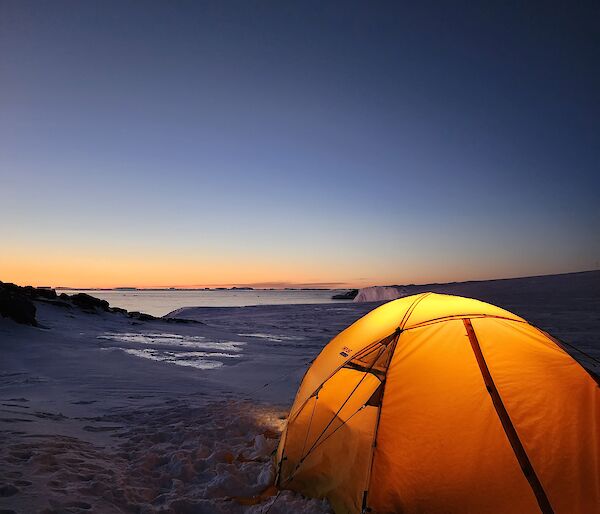 A small yellow tent with a glowing light inside is located in the foreground of the picture. It is set up on snow covered ground and in the distance is a glimpse of the water, and distant icebergs. The sky is glowing orange and blue in the twilight.