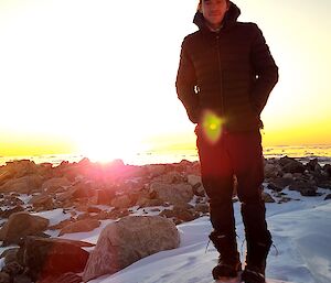 A man is standing on a small rocky hill that is partially covered in snow. He has his hands in his pockets and is facing the camera, as the sun sets in the background.