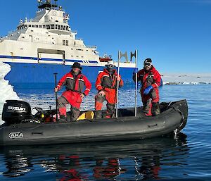 Three men in orange survival suits are standing in a small inflatable boat with long boat hooks in their hands. In the background there is a large blue and white vessel that is providing diesel fuel to the station. There is also a small iceberg to the left of the picture, in between the two boats.