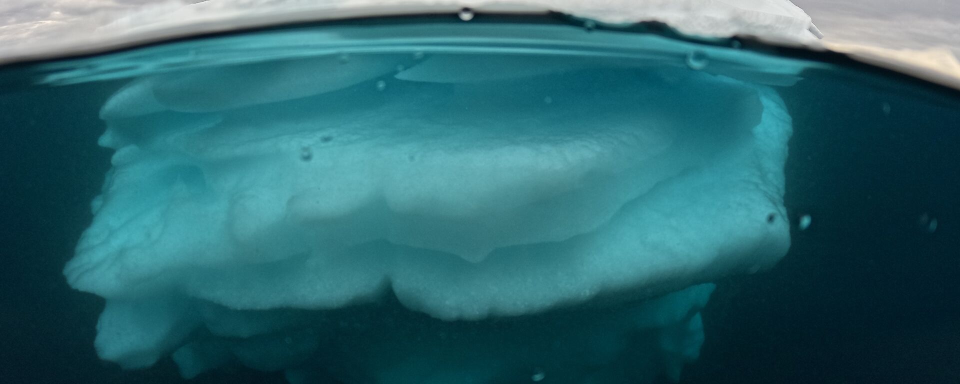 This photo is a cross section of an iceberg. The majority of the white chunk of iceberg is below the waterline and refracting the blueish colour of the water. There is a thin smooth section of ice above the waterline. The sky is grey and cloudy.