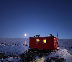 A dark red shipping container of 8m length sits atop a small rocky outcrop that is snow covered. There are two small square windows in the container with a yellow glow in them. It is dusk, and the sky is a dark blue, with a full moon rising just above the horizon in the distant background.