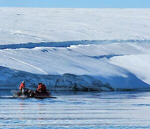 A small inflatable boat with three people dressed in red survival suits is motoring slowly along a calm bay. It the near background is a low flat ice shelf that has a deep crack running along it's length.