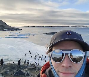 A man stands in the foreground of the photo wearing mirrored sunglasses, a blue baseball cap, and dressed very warmly. In the background of the photo there is an icy shoreline with around 20 penguins gathered on it. The horizon has several small rock islands and the sky is partially covered with clouds. The water out in the bay is calm and flat.