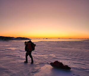 A man with a sled being towed behind him walks from right to left across the photo. He is wearing a large backpack and is dressed warmly. The ground is snow covered and the background sky is a blend of orange, peach, and purple, fading up into the darkening sky. There is a distant patch of glassy flat water in the background, and a low rocky outcrop to the left of the water.