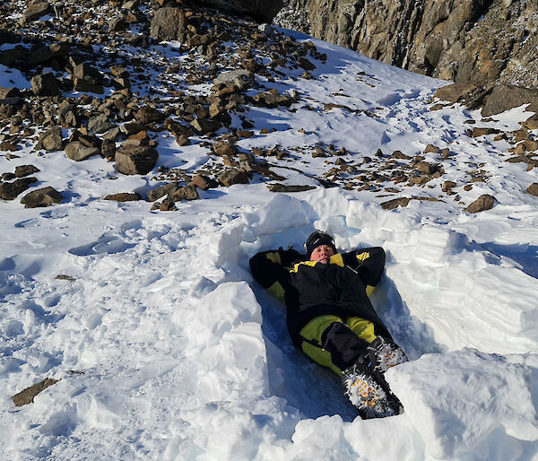 A man is laying in a dug out area in a snowy and rocky landscape