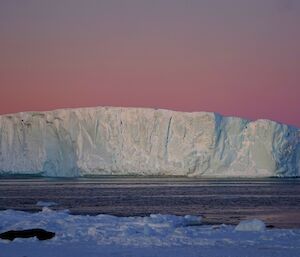 An iceberg is silhouetted against a rose coloured sunset