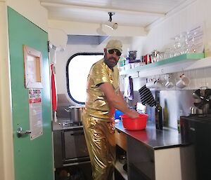 A man is in a small kitchen wearing a gold coloured suit and hat