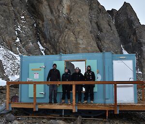 Four people are standing on the deck of a hut with a rocky landscape behind it