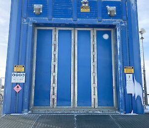 A large blue industrial shed with a blue door with four hinged panels. The door takes up almost the entire face of the building.