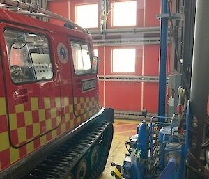 A red Hägglunds fire truck, inside a large industrial shed, facing a large red metal door that goes all the way to the roof