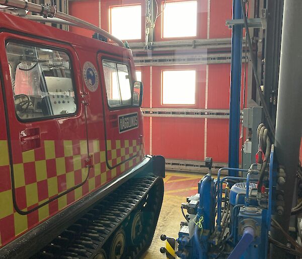 A red Hägglunds fire truck, inside a large industrial shed, facing a large red metal door that goes all the way to the roof