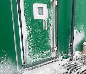A green door, partially covered in snow, providing access to a green building.