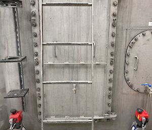A grey metal door with rivets all around the edges leading to the inside of a sewerage tank. There is a pipe underneath the door with a sticker saying "sludge".