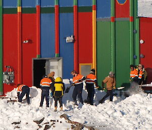 A group of nine people are helping move snow from the front of a building