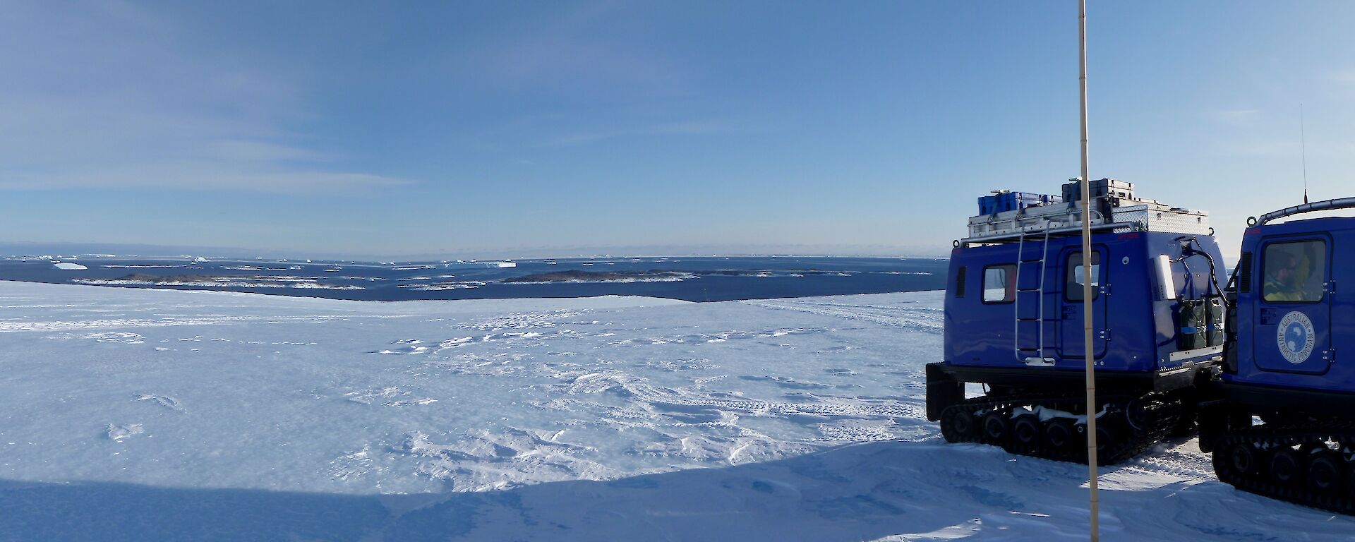 A blue Hägglunds vehicle is on ice with ocean visible in the distance