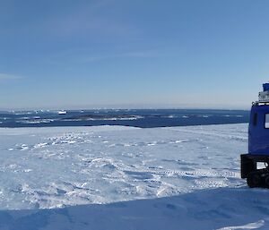 A blue Hägglunds vehicle is on ice with ocean visible in the distance