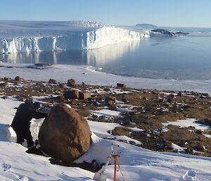 A man is in a rocky, snow covered area with ice cliffs and sea in the background