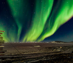 A swirling bright green ribbon of light dances vertically in four strands into the night sky in front of a sign-post (with many signs pointing in different directions) with the sea-ice in the background.
