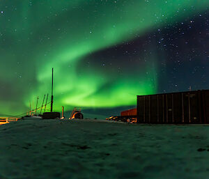 A swirling bright green ribbon of light dances across the night sky over the top of a sea-container, a bridge and a blizz line, at Davis Station