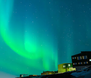 A swirling bright green ribbon of light dances across the night sky over the top of two buildings that are part of Davis Station