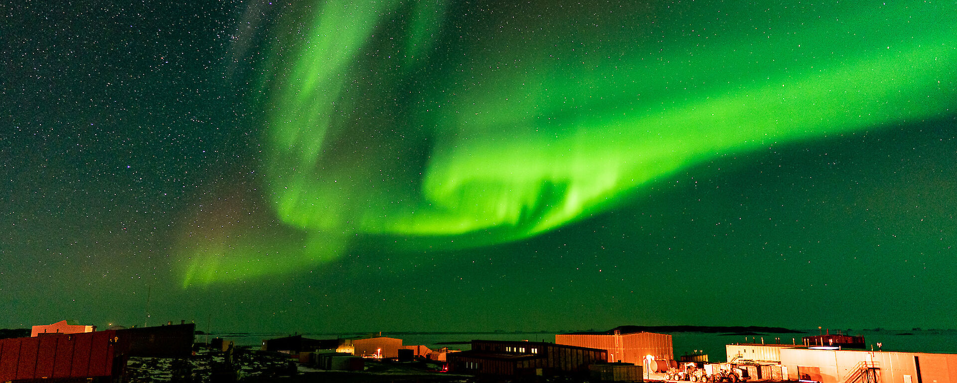 A swirling bright green ribbon of light dances across the night sky over the top of the buildings that make-up Davis Station