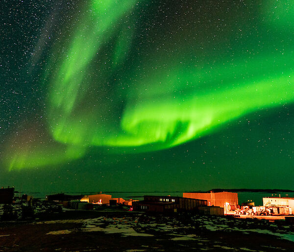 A swirling bright green ribbon of light dances across the night sky over the top of the buildings that make-up Davis Station