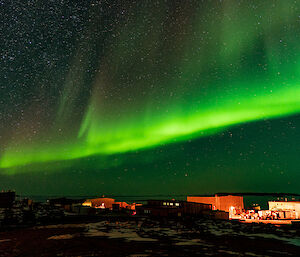 A swirling bright green ribbon of light dances across the night sky over the top of the buildings that make-up Davis Station