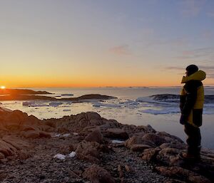 The foreground of this photo is a rocky hill, with sea ice and several other low outcrops of rock stretching into the distance. The sky is a pale blue, with barely any cloud, and a band of dusky orange light dividing the sky and land. The sun can be seen half set below the horizon to the very left of frame. Dr Sophie is standing on top of the hill, to the right of frame dressed in warm yellow and black survival clothes. She is facing the setting sun.