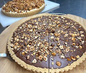 This photo shows large a round chocolate dessert tart, covered in crushed nuts, sitting on a wooden serving platter. In the background is another dessert tart.