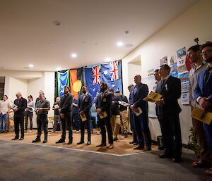 A line of approximately 20 people, all dressed formally, stand facing the left of frame, most with their hands clasped together in front of them and holding small paper programmes. In the background are the flags of the Torres Strait Islands, Aboriginal, New Zealand, and Australia, hanging side-by-side from the ceiling.