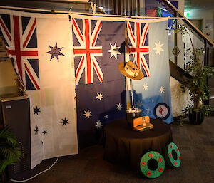 The cenotaph for our inside service. It is an Army slouch hat, resting atop an ice-pick. The pick is inserted into a black plant pot filled with white stones, and is sitting on a low round table with a black table cloth covering it. There is a small glass container with sand from the beach at Gallipoli sitting in front of the plant pot. On the ground in front of the table are two green circular wreaths, with red poppies on them. The background is the White Australian Navy Flag, the Australian Flag, and the light blue Airforce Flag hanging side-by-side from the stairs above.