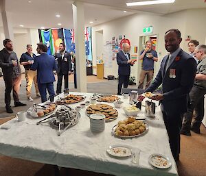 A large square table with a white tablecloth occupies the foreground of this photo. The table is covered in platters of biscuits, lamingtons, and pies. There is a man in a dark blue suit to the right of table, placing food from the table onto his plate. Two groups of four people each are in the background chatting, all are dressed formally.