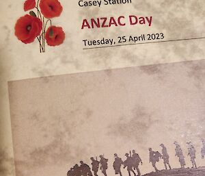 This photo is a close up shot of the official programme front cover. The paper is a faded yellow parchment style, with the words "Casey Station, ANZAC Day, Tuesday 25 April 2023" next to a picture of four red poppies. Below these words is a photograph from World War One of a line of soldiers in silhouette, marching along a ridge line.