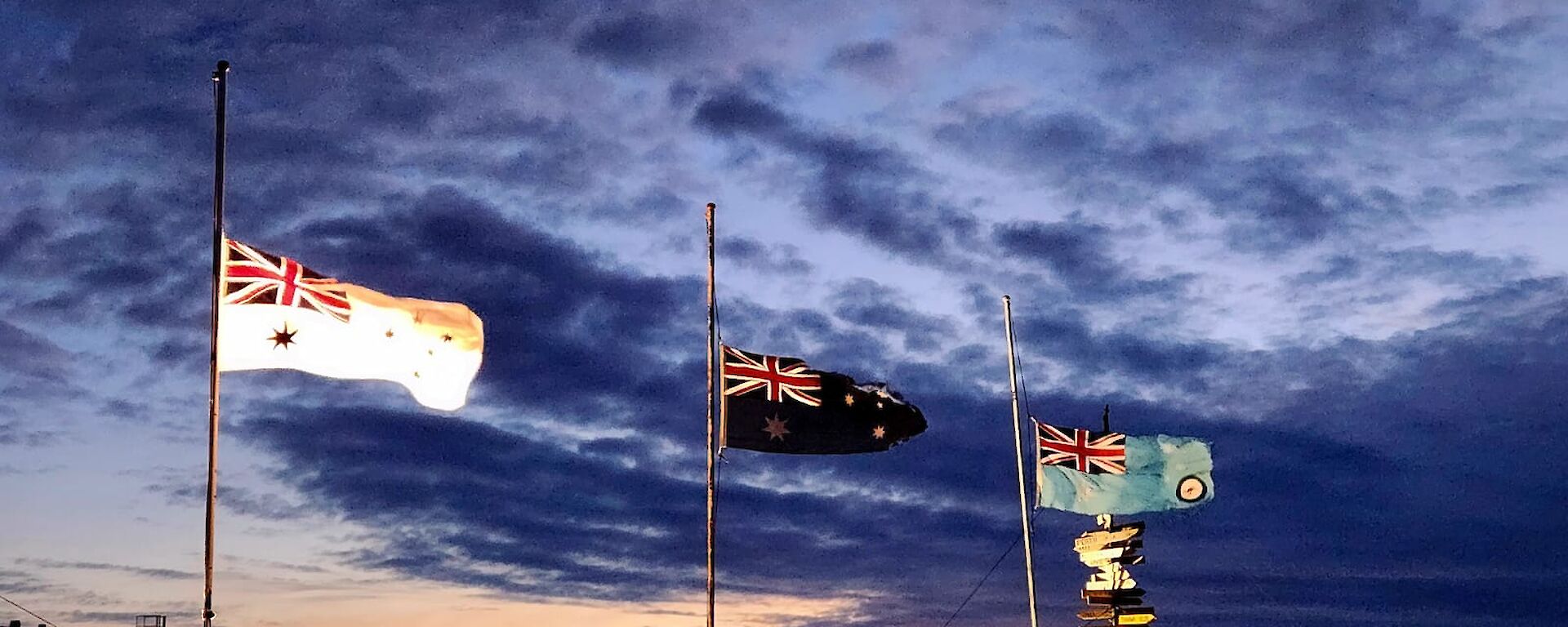 Three flag poles occupy the main part of the photo, with flags at half mast, and a light orange sunset in the background. The flags are all Australian, with the left one being white for the Navy, the centre one is the dark blue Australian Flag, and the right hand flag is light blue for the Airforce. The flag poles are mounted on a snow covered rocky hill and the sky is partly cloudy.
