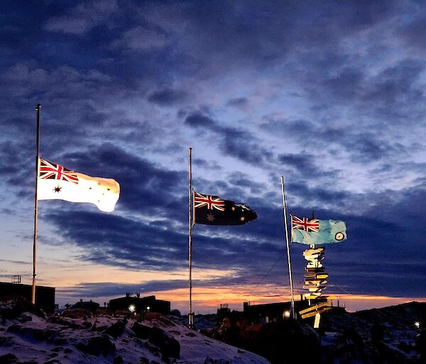 Three flag poles occupy the main part of the photo, with flags at half mast, and a light orange sunset in the background. The flags are all Australian, with the left one being white for the Navy, the centre one is the dark blue Australian Flag, and the right hand flag is light blue for the Airforce. The flag poles are mounted on a snow covered rocky hill and the sky is partly cloudy.